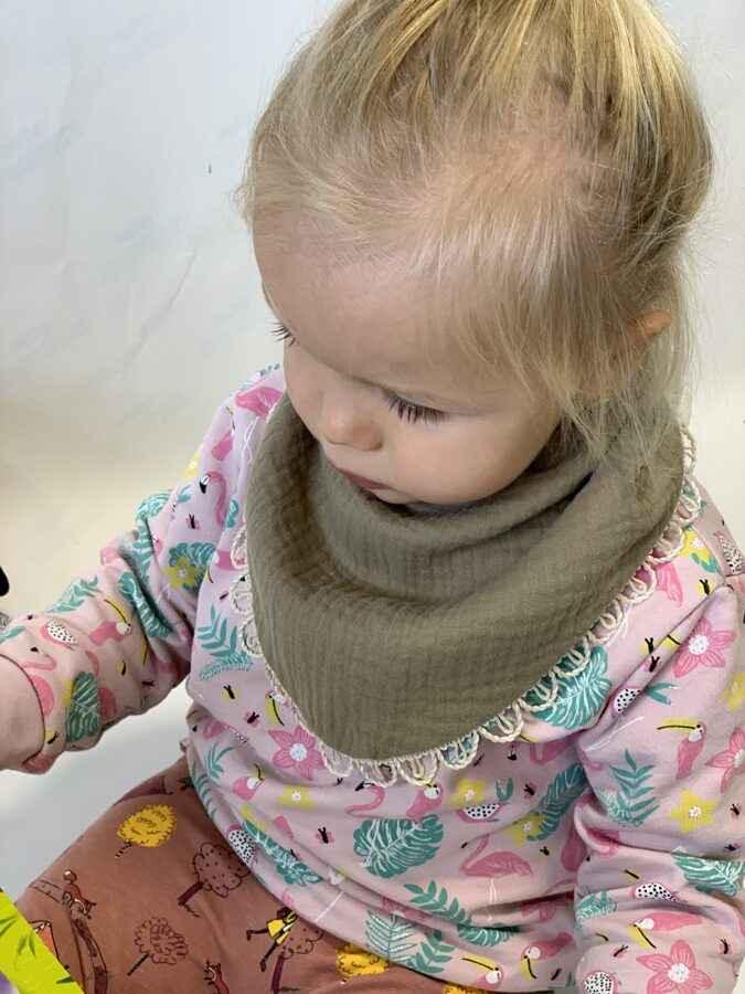 Toddler wearing a pink animal print top and beige cotton bib scarf, sitting indoors